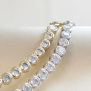 Two tennis bracelets with round clear gemstones, one in yellow gold and one in silver, displayed on a cream satin jewelry stand.