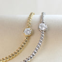 Two beaded bracelet-style necklaces displayed on a cream fabric surface, one in yellow gold and one in silver, each featuring a round clear gemstone in a prong setting.
