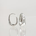 A pair of polished silver hoop earrings with a textured, organic wavy surface, displayed on a reflective light gray surface.