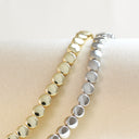 Two beaded bracelets with faceted round beads, one in gold and one in silver, displayed on a beige fabric surface.