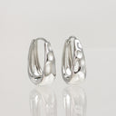 A pair of polished silver huggie hoop earrings with a textured, organic hammered surface, displayed on a reflective white surface against a light gray background.