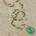 A gold chain necklace with small green beads and a large teardrop-shaped turquoise pendant featuring a green stone in the center, displayed on a textured beige stone surface.