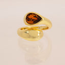 A gold wrap-style ring featuring a teardrop-shaped amber gemstone, displayed on a clean, light beige reflective surface.