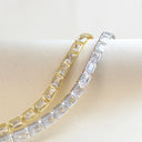 Two tennis-style bracelets with rectangular clear stones, one in yellow gold and one in silver, displayed on a cream fabric surface.