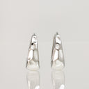 A pair of textured, hammered silver hoop earrings with a tapered, organic shape, displayed on a reflective light gray surface against a neutral light gray background.