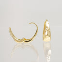 A pair of gold hoop earrings shown on a reflective light gray surface, with one earring facing forward and the other angled to the side to show the back.