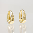 A pair of gold huggie hoop earrings with a textured, organic wavy surface, displayed on a reflective light gray surface.