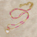 A double-strand gold chain necklace featuring bright pink faceted beads and a gold heart-shaped pendant with a clear crystal, displayed on a light beige marble surface.