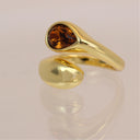 A gold ring with a teardrop-shaped amber gemstone, displayed on a reflective light pink surface.