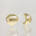 A pair of textured gold hoop earrings with a ribbed, hammered finish, shown on a reflective light gray surface. One earring is shown from the front, displaying its spherical shape and vertical texture, while the other is shown from the side, displaying its curved shape and clear plastic backing.