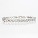A silver tennis bracelet with round-cut clear gemstones in a bezel setting, displayed on a reflective white surface.