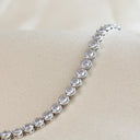 A silver tennis bracelet with round clear gemstones in a row, displayed on a beige fabric jewelry stand.