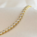 A gold tennis bracelet with round clear gemstones, displayed on a beige satin fabric surface.
