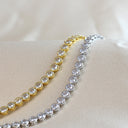 Two tennis-style bracelets with round clear stones, one in yellow gold and one in silver, displayed on a beige satin surface.