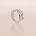 A polished silver ring with three parallel, slightly curved bands, displayed on a reflective cream-colored surface.