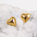 Two shiny gold heart-shaped stud earrings resting on white salt crystals against a light gray background.