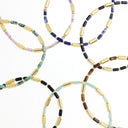 Multiple beaded jewelry strands arranged in overlapping loops on a white background, featuring metallic gold cylindrical beads alternating with stones in blue, green, black, brown, and pale lavender.