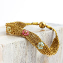 A gold chain-link woven bracelet with three small floral charms in pink, mint green, and white, resting on a rustic white wooden block.