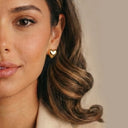 A close-up of a woman wearing a small gold heart-shaped stud earring, showing her ear, hair, and part of her face against a soft beige background.