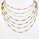 A set of six layered beaded necklaces arranged in a fan shape on a plain white background. Each necklace features a gold-tone chain with small, round beads in different colors including pink, light blue, dark blue, green, red, black, and white.