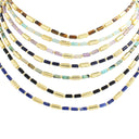 Five layered beaded necklaces arranged in a fan shape on a plain white background. The necklaces feature metallic gold cylindrical beads alternating with various colored stone beads including blue, green, purple, brown, and black.