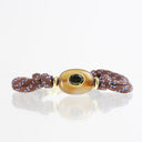 A handmade braided bracelet featuring a central polished oval stone with a green center, flanked by gold-toned spacers and a knotted brown cord with blue and white speckled accents, shown on a reflective white surface.