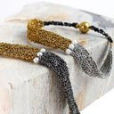 Two woven chain bracelets with black cord and white pearl accents, displayed on a light stone block. One bracelet is gold-toned and the other is silver-toned, both featuring a polished gold bead accent.
