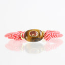 A red and white patterned cord bracelet featuring a central brown tiger's eye bead with a round red gemstone in a gold bezel setting, displayed on a reflective white surface.