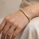 A close-up of a person's hand wearing a delicate gold chain bracelet with polished rectangular links, resting on a soft beige fabric surface.