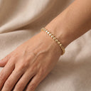 A delicate gold chain bracelet with small, polished heart-shaped links, worn on a woman's wrist against a soft beige fabric background.