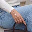 A close-up of a person's hand wearing a delicate gold chain hand bracelet with two small round crystal charms, styled with a white ribbed knit sweater and light blue jeans.