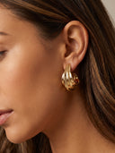A close-up of a woman's ear wearing a gold crescent-shaped hoop earring with a textured, layered design.