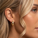 A close-up of a model's ear wearing a small gold huggie hoop earring adorned with colorful enamel dots in green, pink, and blue.