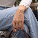 A close-up of a person's hand and leg wearing a delicate gold chain hand bracelet with small clear stones, paired with light blue jeans and a white sweater.