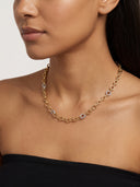 A close-up of a model wearing a gold chain necklace with small oval links and a few clear gemstone accents. The model is wearing a black strapless top against a plain light background.