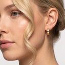 Close-up of a woman's ear wearing a small gold hoop earring with a clear gemstone, shown against a light gray background.