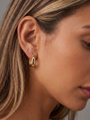 A close-up of a woman's ear wearing a small gold hoop earring with a polished, reflective finish.