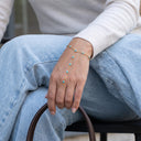 A close-up lifestyle shot of a person's hand wearing a delicate gold chain hand bracelet with small turquoise blue stones, paired with a matching gold ring, while sitting in light blue jeans and a white sweater.