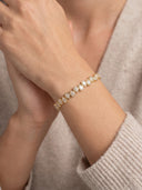 A close-up of a delicate gold bracelet with small clear stones, worn on a person's wrist. The person is wearing a beige knit sweater, and their hands are clasped in front.