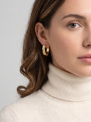 A close-up of a woman wearing a chunky, textured gold hoop earring, paired with a cream-colored ribbed turtleneck sweater against a neutral light gray background.