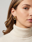 A close-up of a woman wearing a pair of gold double-ring hoop earrings, styled with a cream ribbed turtleneck sweater against a neutral light gray background.
