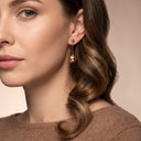 A close-up of a woman wearing a gold dangle earring with a small bead drop, shown against a soft beige background.