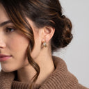 A close-up of a woman wearing a small gold hoop earring with clear gemstones, shown from the side against a light gray background.
