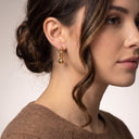 A close-up side profile of a woman wearing a small gold dangle earring with three stacked polished beads, against a clean light gray background.