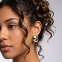 A close-up of a woman wearing a large, round, polished gold earring, shown from the side against a neutral light gray background.