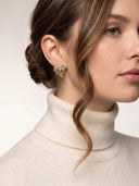 A close-up of a woman wearing a textured gold heart-shaped stud earring, paired with a cream ribbed turtleneck sweater against a light neutral background.
