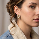 A close-up of a woman wearing a textured gold crescent-shaped hoop earring, styled with a denim jacket featuring a cream sherpa collar.