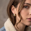 A close-up of a woman wearing a textured, organic-shaped gold earring with a small central clear stone, styled with a denim jacket featuring a cream sherpa collar.
