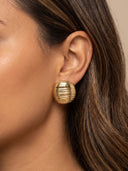 A close-up of a woman's ear wearing a textured, ribbed gold stud earring against a warm beige background.