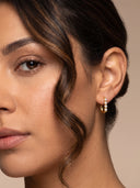 A close-up of a woman's ear wearing a small gold huggie hoop earring adorned with a row of white pearls, set against a soft beige background.